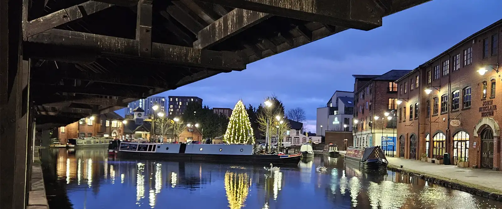 The view from Spirit Bridge Studio at the Coventry Canal Warehouse, looking across the canal at dusk. The water reflects the lights from canal boats, lamp posts, and a large, illuminated Christmas tree in the canal square.