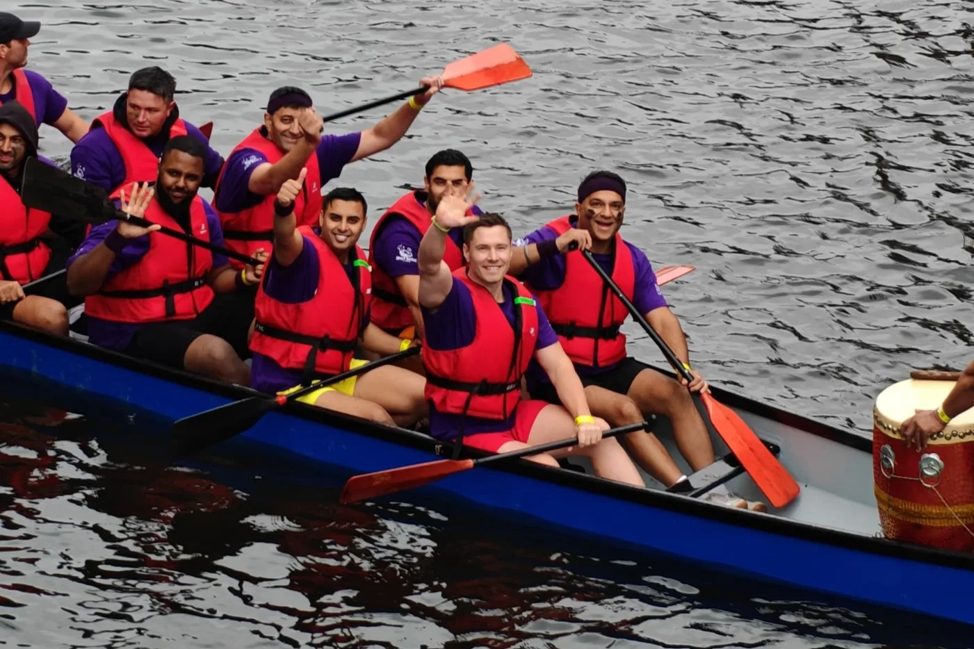 The Spirit Bridge Studio team, "Team Bucky," smiling and waving to the camera from a blue dragon boat during the 2025 Dragon Boat Race at the Mailbox canal in Birmingham. The crew is wearing matching purple Bucky t-shirts and red life vests, celebrating together on the water.